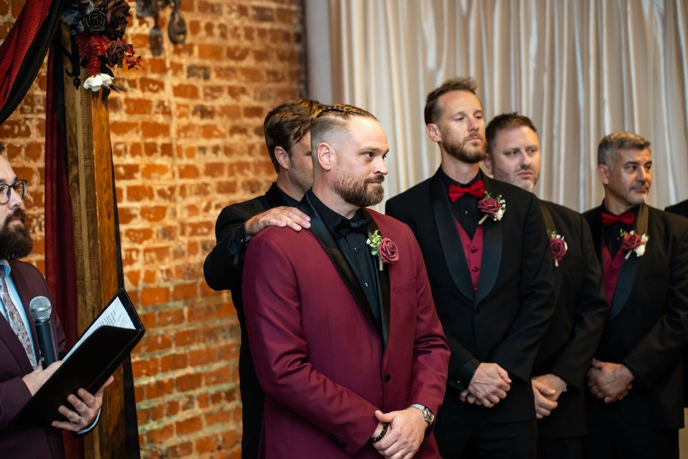 Groomsmen in dark suits with red boutonnieres stand near a groom in a burgundy jacket during a wedding ceremony in front of a brick wall.