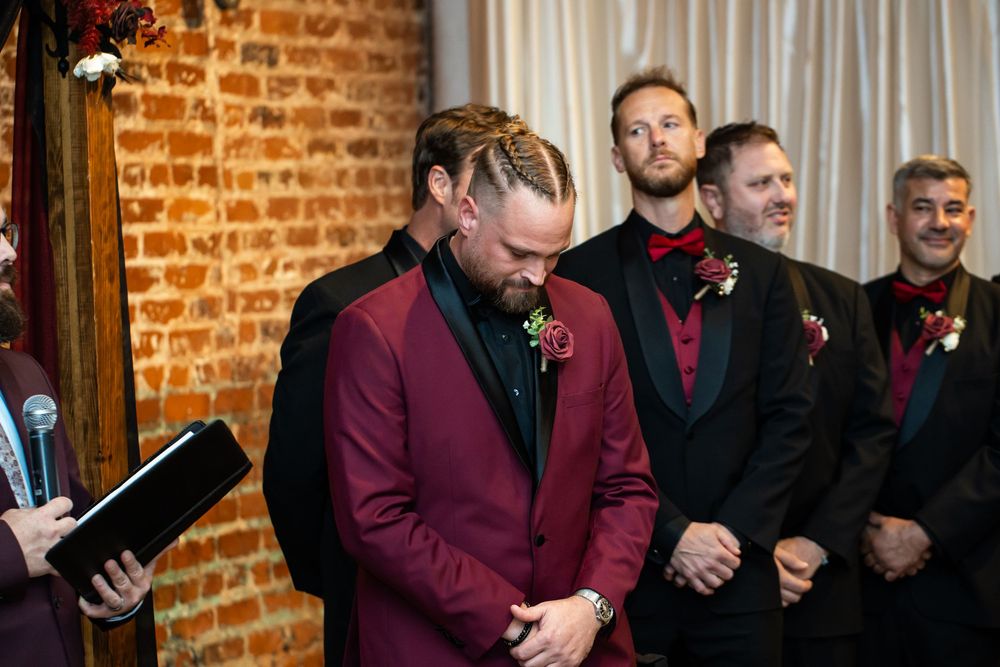 Groom in a burgundy velvet tuxedo with a floral boutonniere stands emotionally at the altar as groomsmen in matching attire look on.