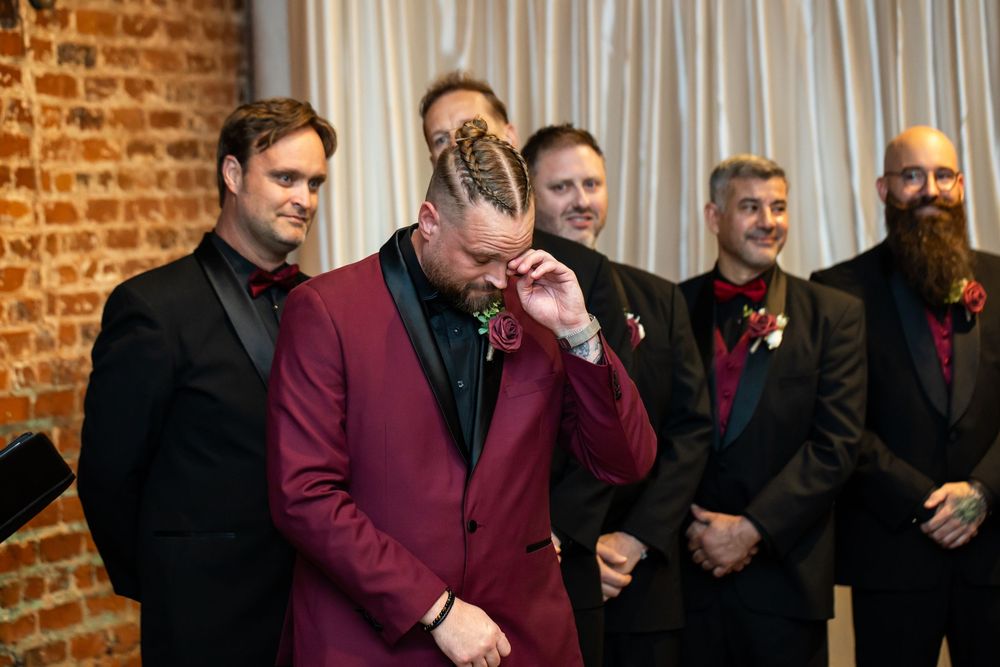 Emotional groom in a burgundy suit wipes his eye at the altar while groomsmen in black suits and boutonnieres smile beside him.