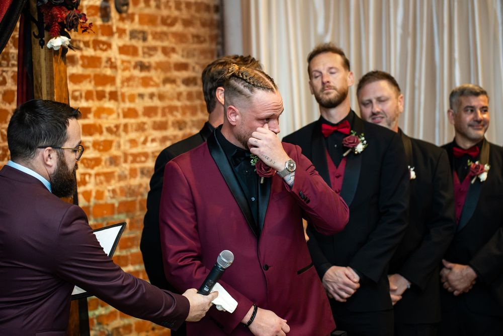 Emotional groom in a burgundy suit wipes tears during his wedding ceremony while groomsmen in dark suits smile beside a brick wall.