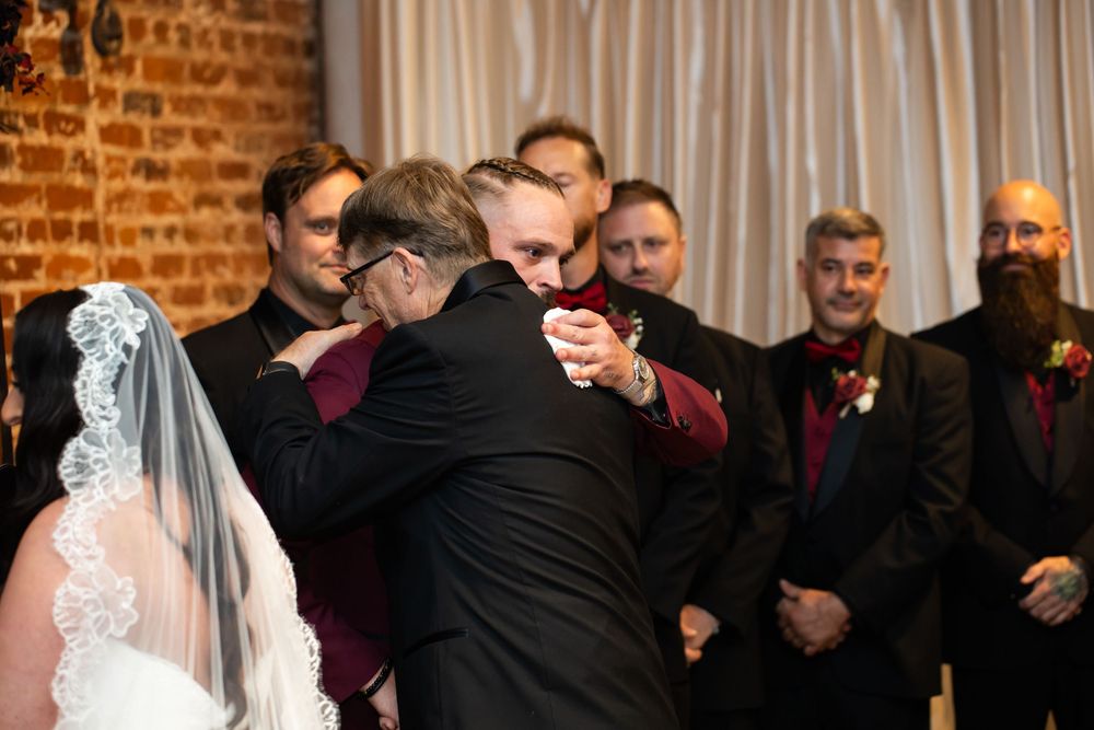 Groomsmen in dark suits watch as a man in black embraces a veiled bride during a wedding ceremony in front of a brick wall.