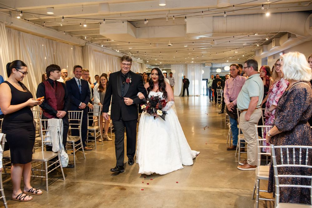 Bride in a white gown holding a bouquet walks down the aisle on the arm of a man in a dark suit as seated wedding guests look on.