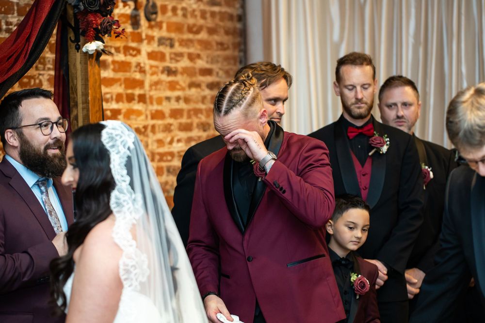 A groom in a burgundy velvet blazer wipes his eyes during a wedding ceremony as the veiled bride and groomsmen look on.
