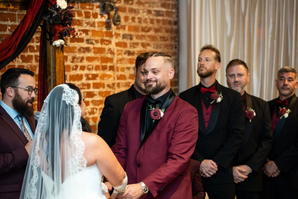 Groom in a burgundy suit holds hands with a veiled bride at a rustic brick-walled wedding ceremony as groomsmen in black suits look on.