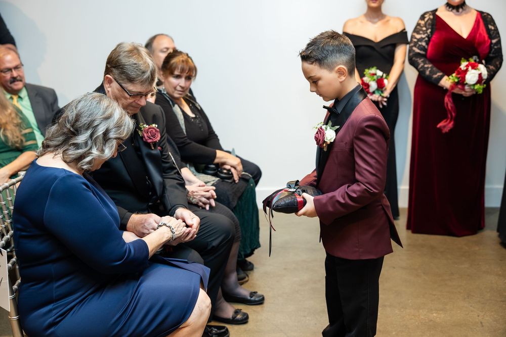Young ring bearer in a burgundy suit holds a small pillow as seated wedding guests in formal attire watch during the ceremony.