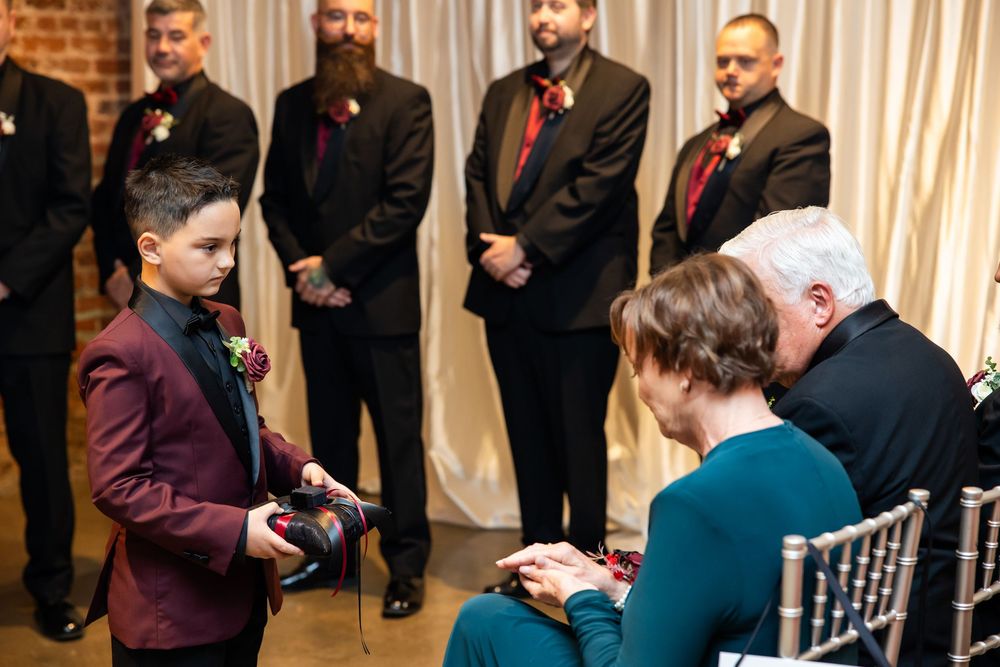 Young ring bearer in a burgundy suit holds a ring pillow at a wedding ceremony while groomsmen in dark suits stand behind and guests watch.