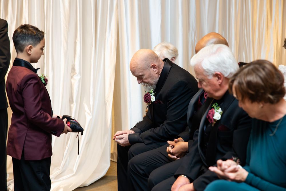 Ring bearer in a burgundy jacket holds a small pillow while seated wedding guests bow their heads in prayer during the ceremony.