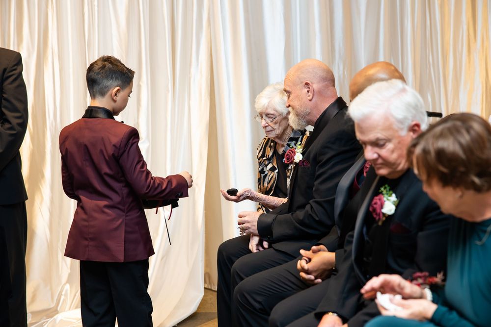 Boy in a burgundy jacket stands before seated elderly guests wearing floral corsages at a ceremony in front of draped white curtains.