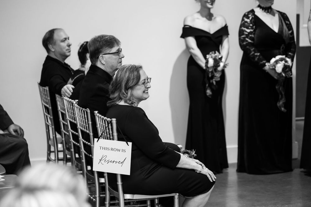 Guests in dark attire sit in chairs beside a reserved sign at a wedding ceremony while bridesmaids in black gowns hold bouquets.
