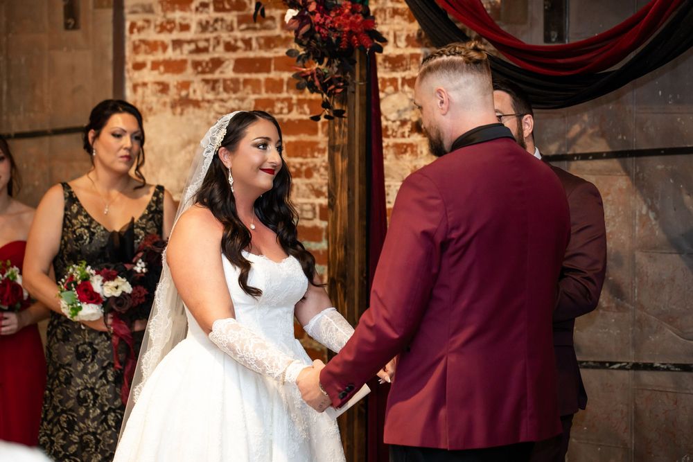 Bride in white lace gown and veil holds hands with groom in burgundy velvet jacket during a wedding ceremony as a bridesmaid watches.