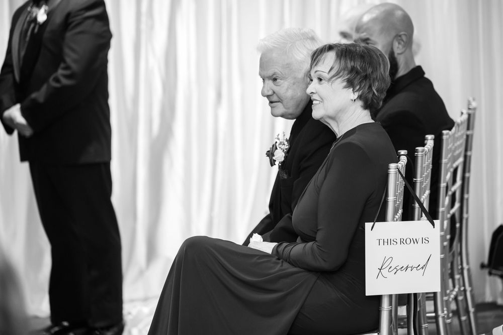 Black and white photo of an older couple seated in the front row at a wedding ceremony beside a chair sign reading This row is reserved.