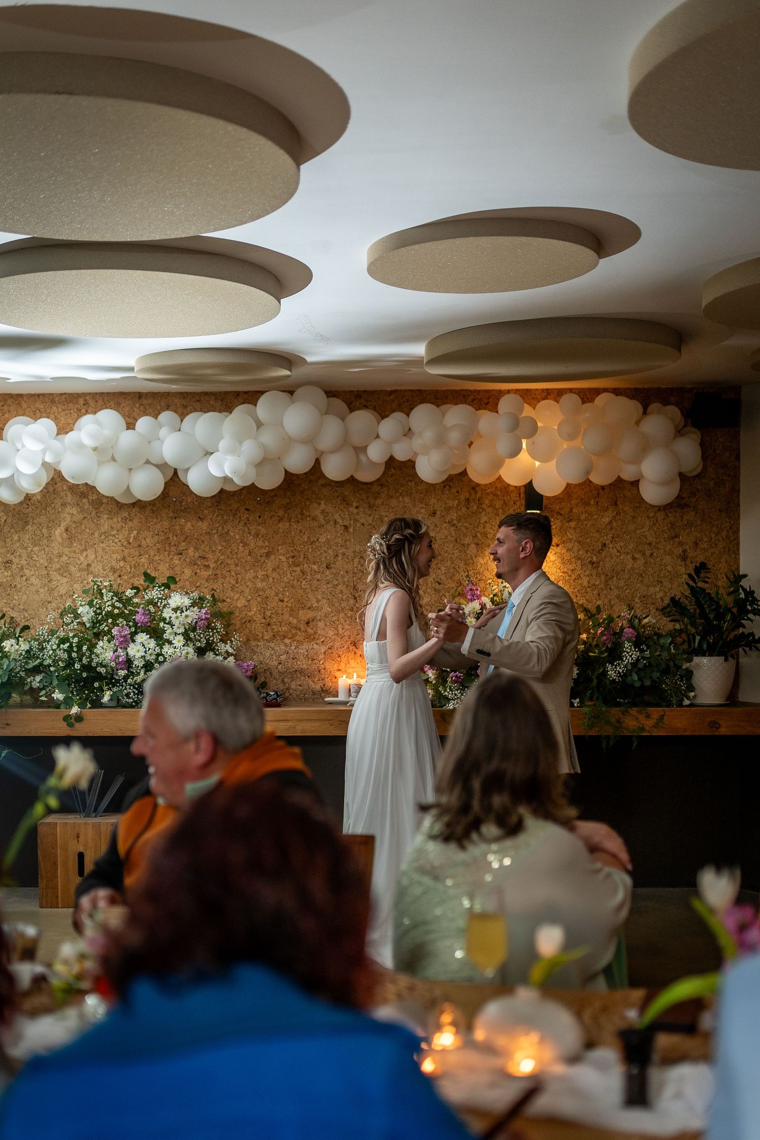 Bride and groom share a toast at their wedding reception, surrounded by white balloon arch and candlelit tables.