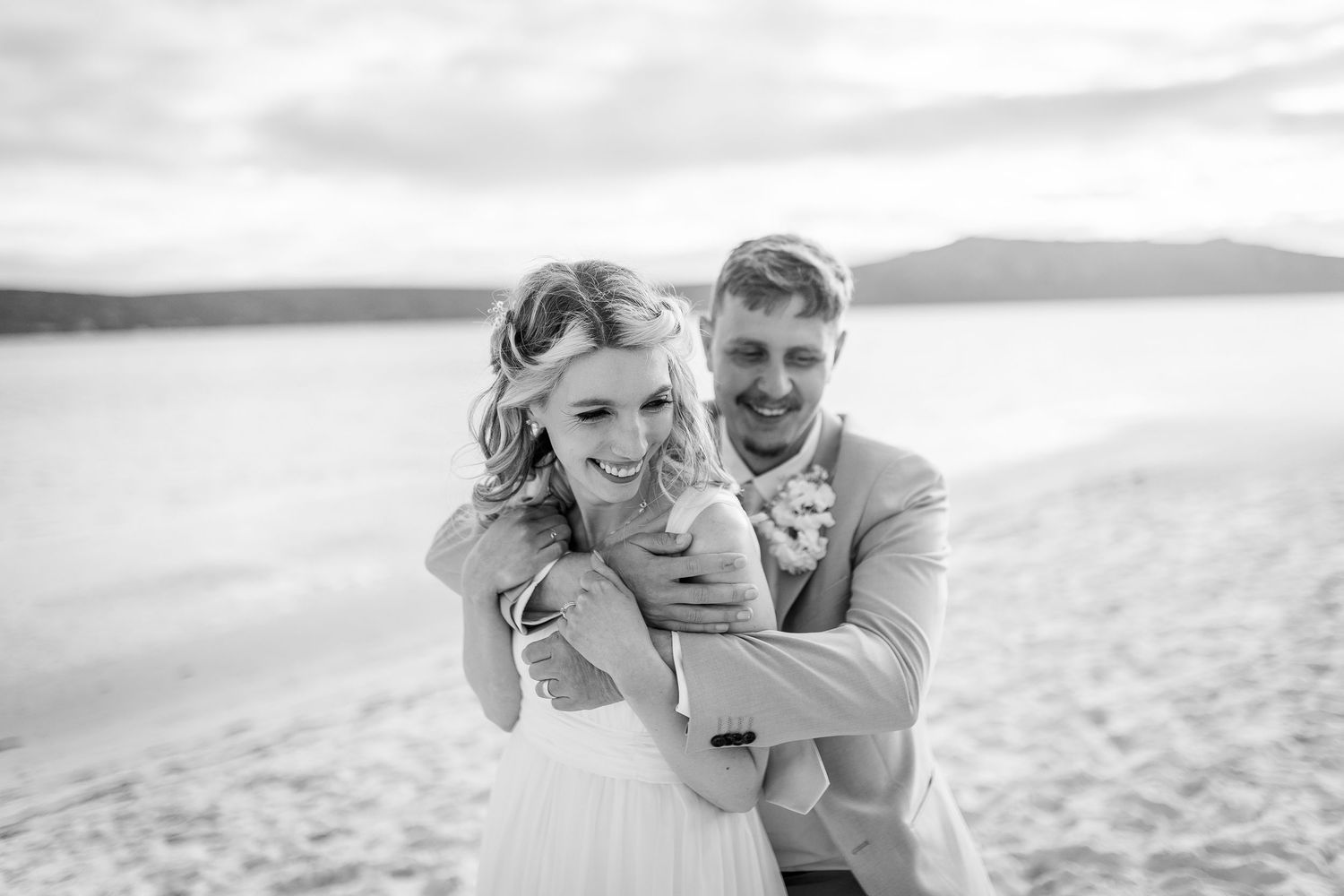 Black and white beach wedding photo of smiling bride and groom embracing on sandy shore with mountains in background.