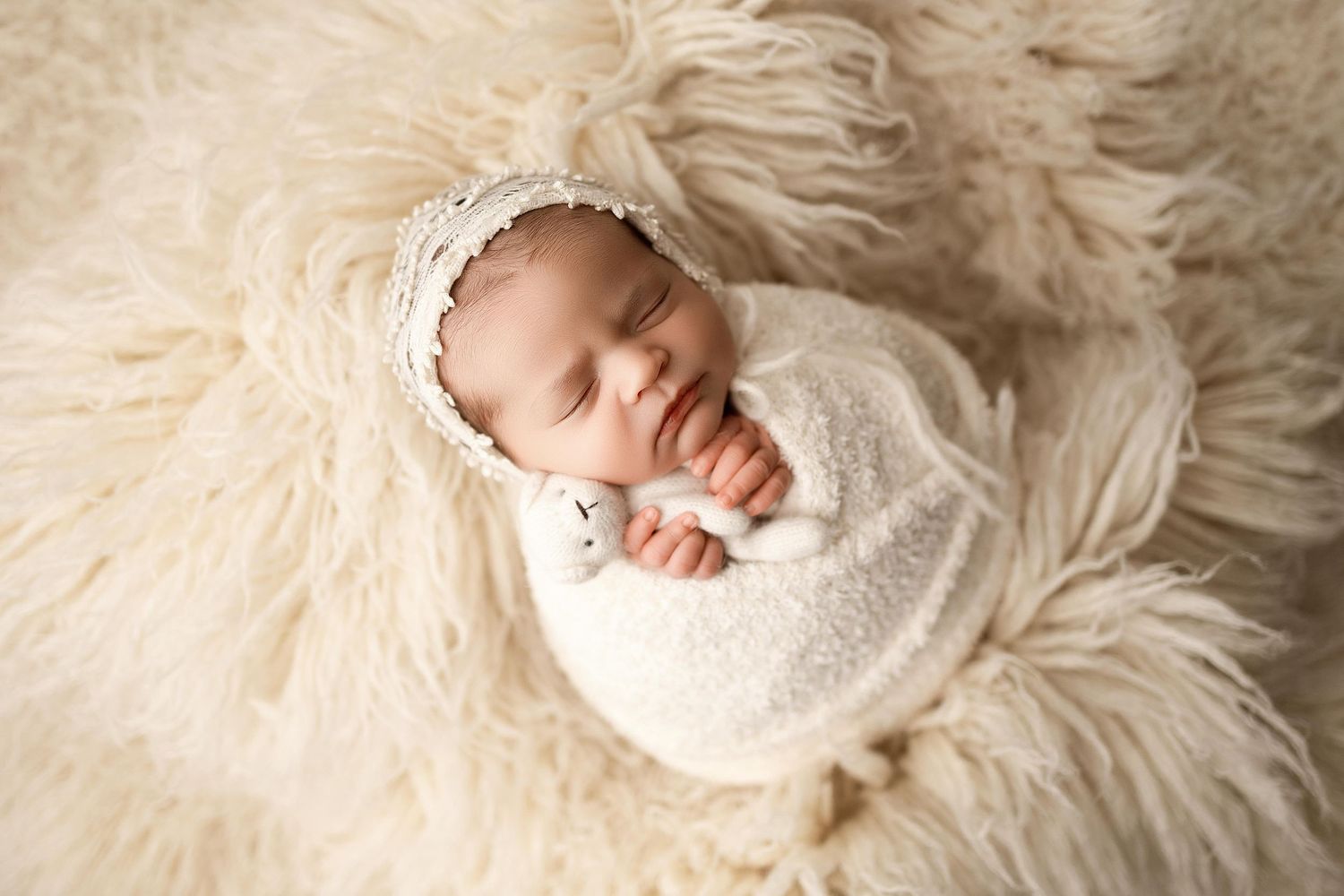 Newborn baby swaddled in cream knit wrap and bonnet, sleeping peacefully on a fluffy white fur rug.
