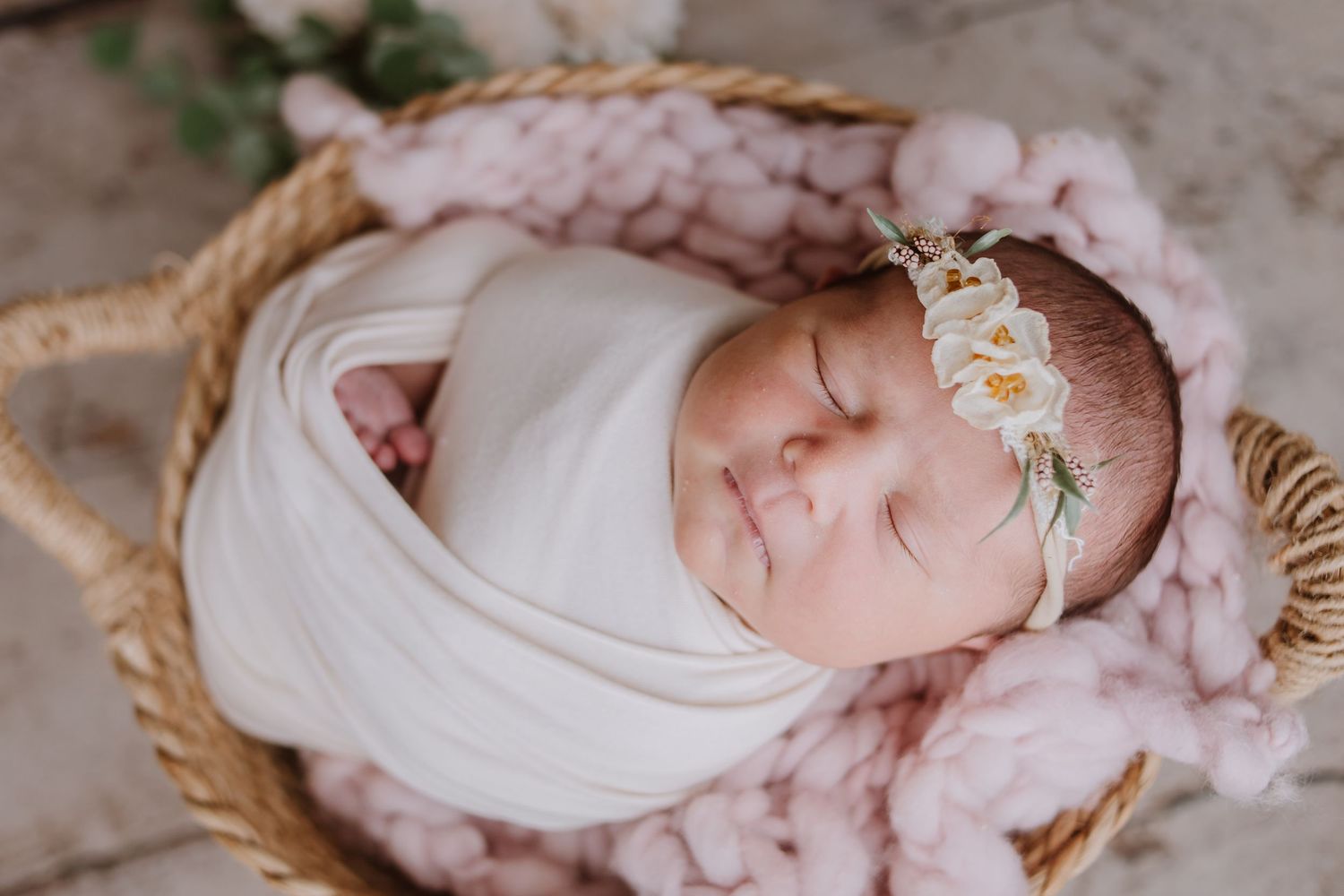Newborn baby swaddled in white wrap sleeping in wicker basket with pink knit blanket and floral headband.