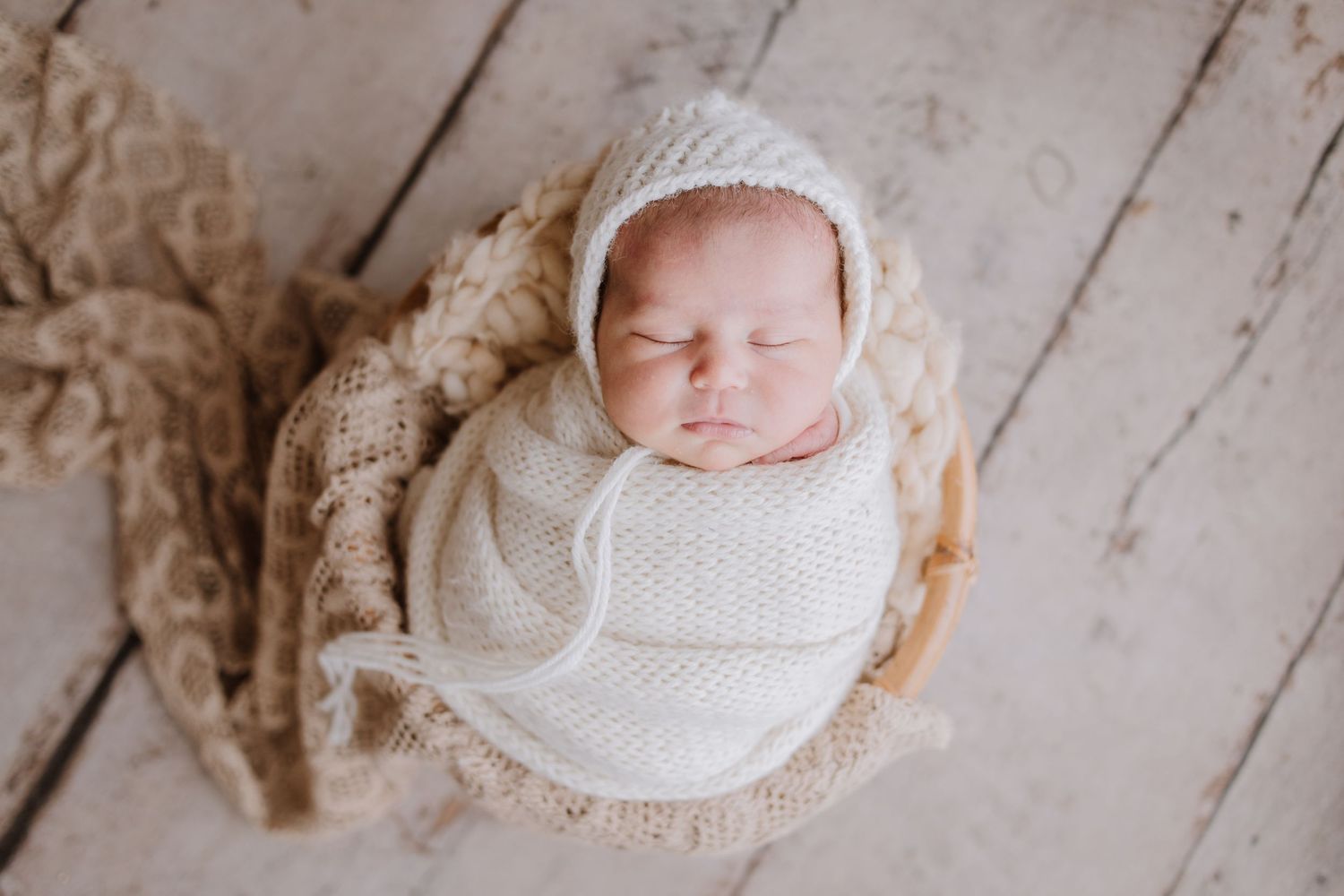 Peaceful newborn baby swaddled in cream knit wrap and bonnet, sleeping in a wicker basket on white wood.
