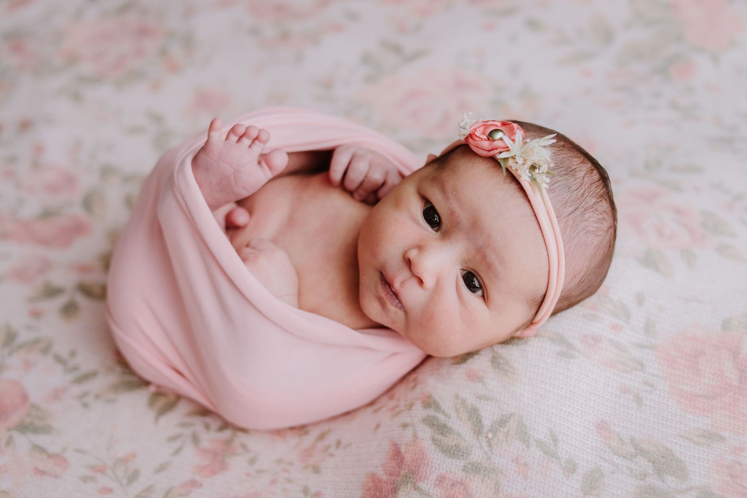 Newborn baby girl wrapped in pink swaddle with floral headband, smiling on a rose-patterned backdrop.