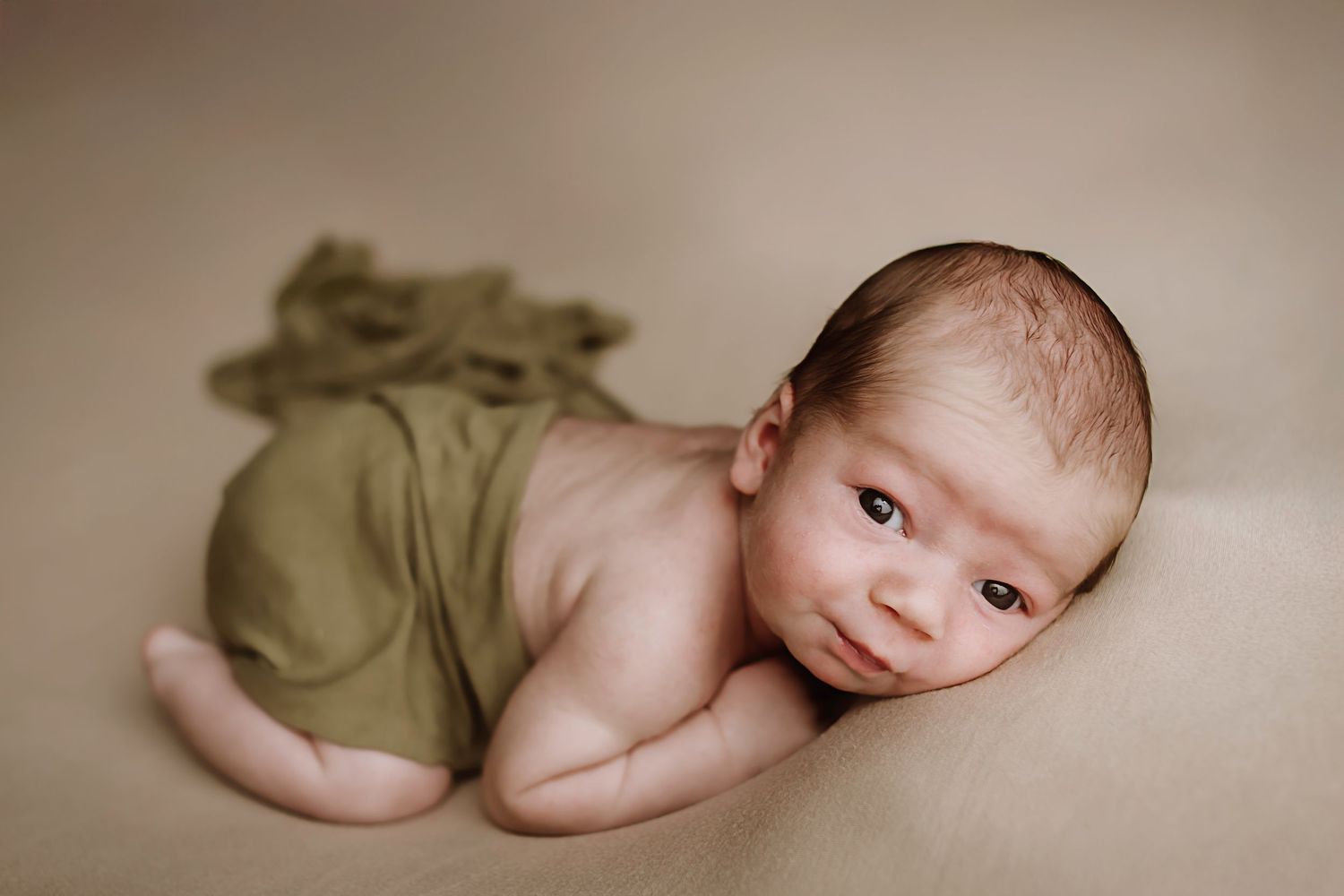 Newborn baby wrapped in olive green cloth lying on beige backdrop in professional newborn photography session.