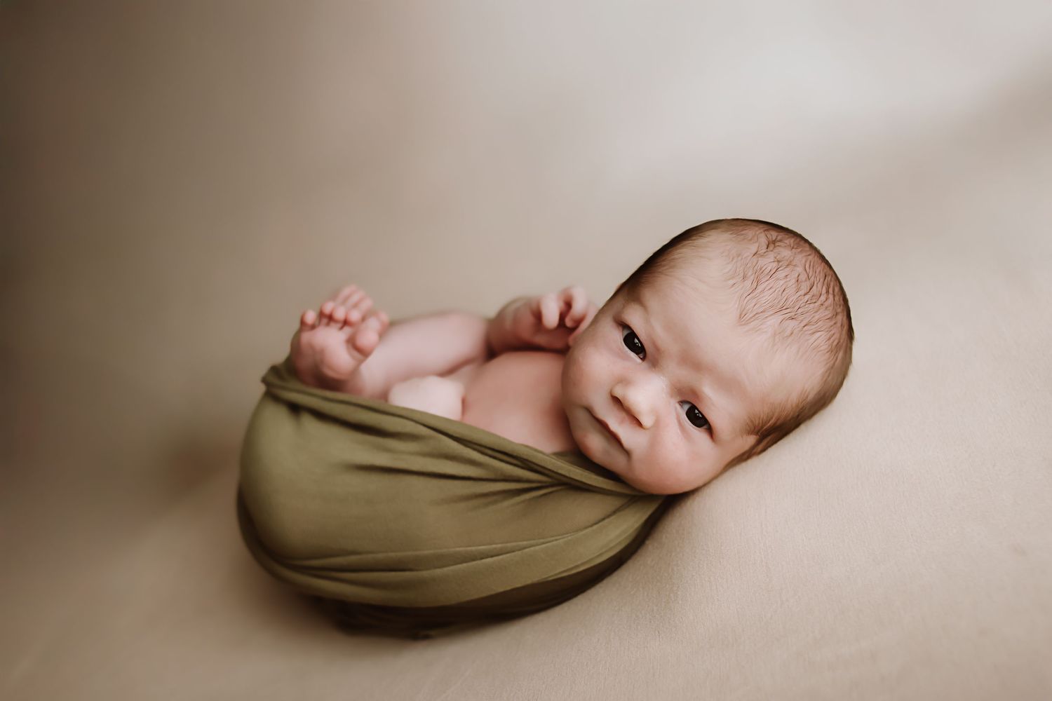 Newborn baby wrapped in olive green swaddle cloth posed on beige backdrop in professional studio photo.