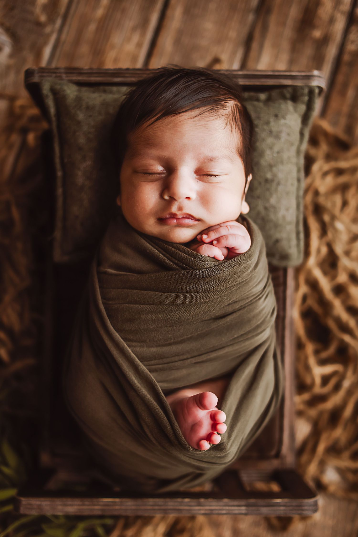 Sleeping newborn baby swaddled in olive green wrap, resting on a small wooden crate with a green pillow.