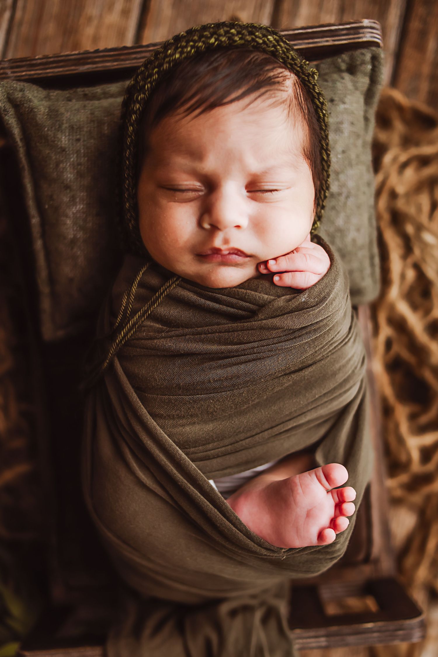 Sleeping newborn baby swaddled in olive green wrap wearing knit bonnet, resting peacefully in rustic wooden box.