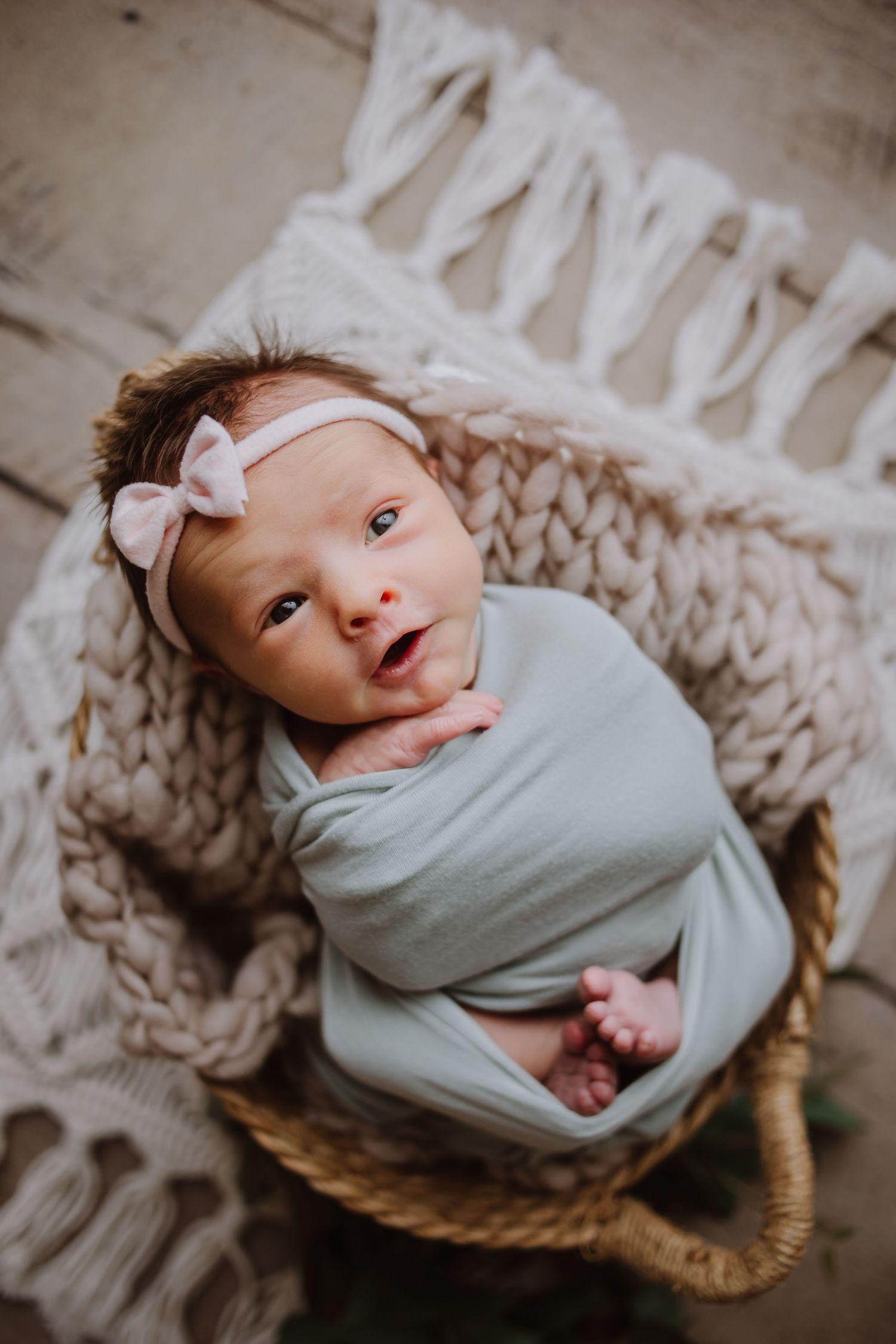 Newborn baby girl swaddled in mint wrap, wearing pink bow headband, resting in wicker basket on knit blanket.