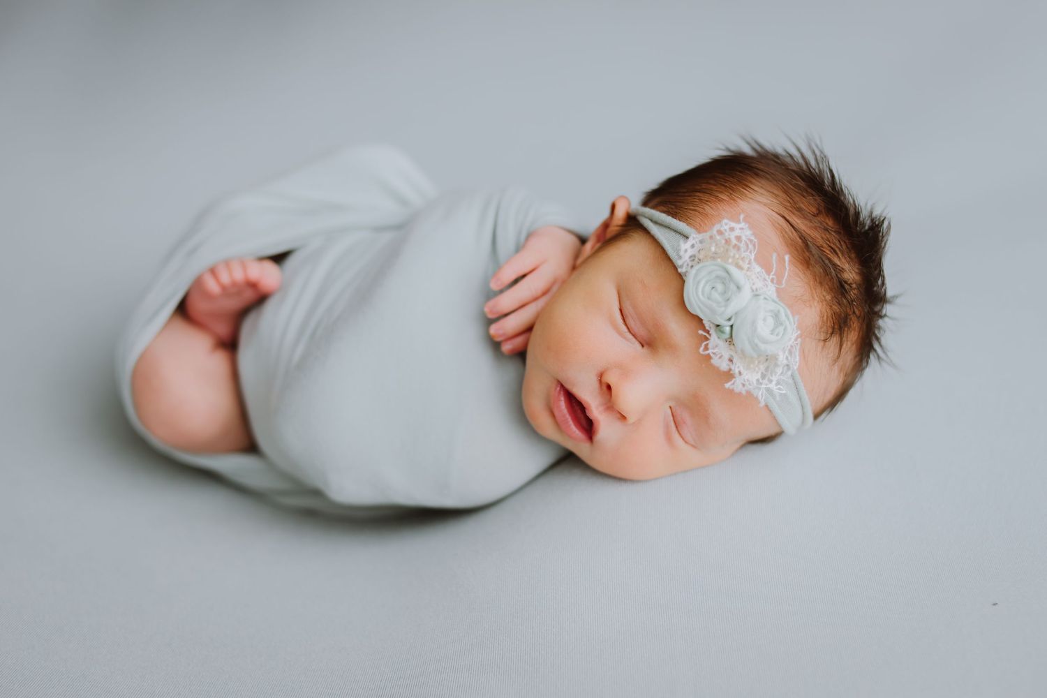 Sleeping newborn baby swaddled in a soft gray wrap wearing a delicate white lace headband on a gray background.