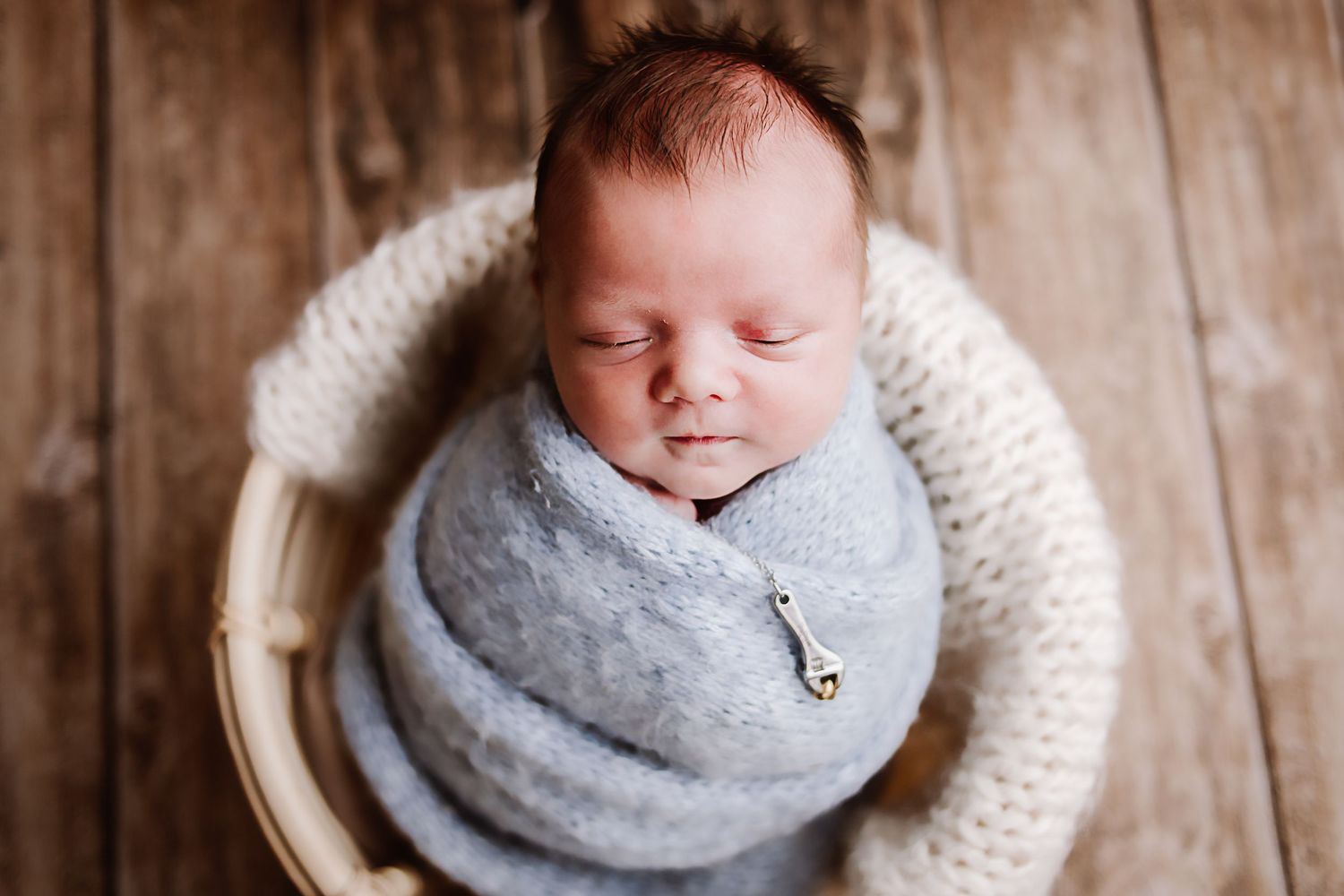 Sleeping newborn baby swaddled in a soft blue wrap, nestled in a wooden bowl with a cream knit blanket.
