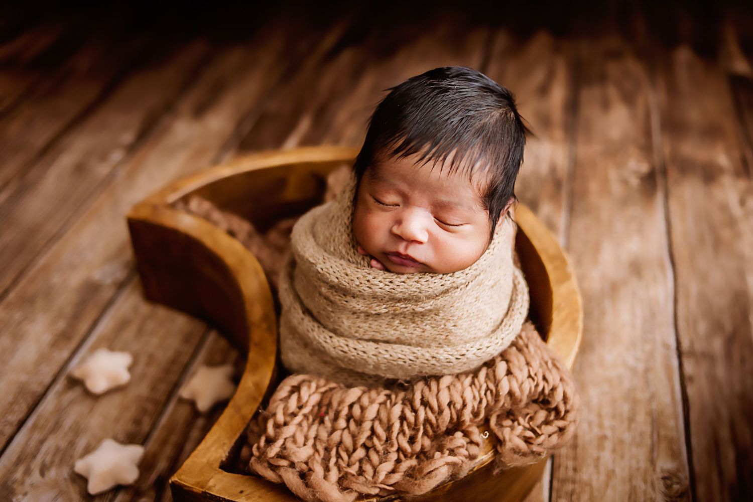 Peaceful newborn baby wrapped in a cozy knit blanket, sleeping in a wooden bowl on a rustic wood floor.