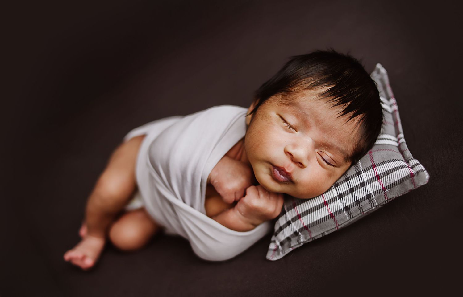 Peaceful newborn baby sleeping wrapped in white swaddle on plaid pillow against dark brown background.