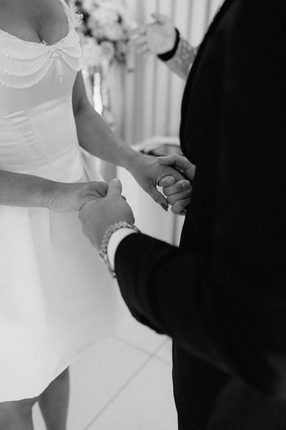 Black and white photo of bride and groom holding hands during wedding ceremony, bride wearing pearl bracelet.