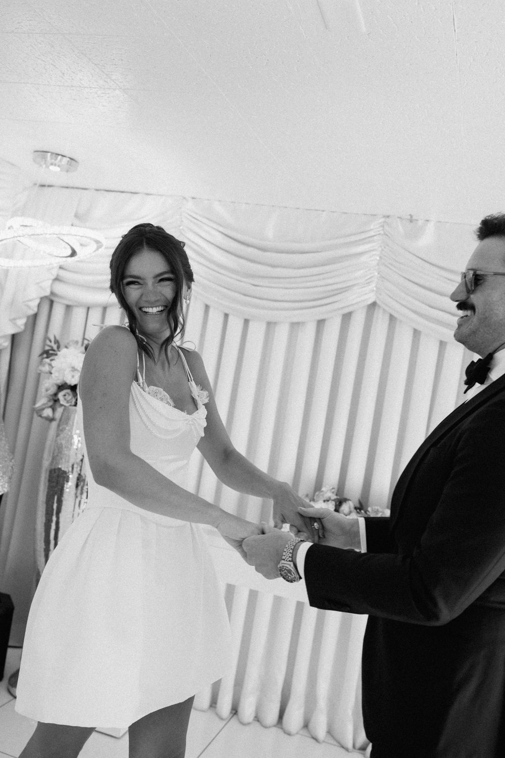 Black and white photo of a smiling bride in a short white dress holding hands with a groom in a tuxedo.