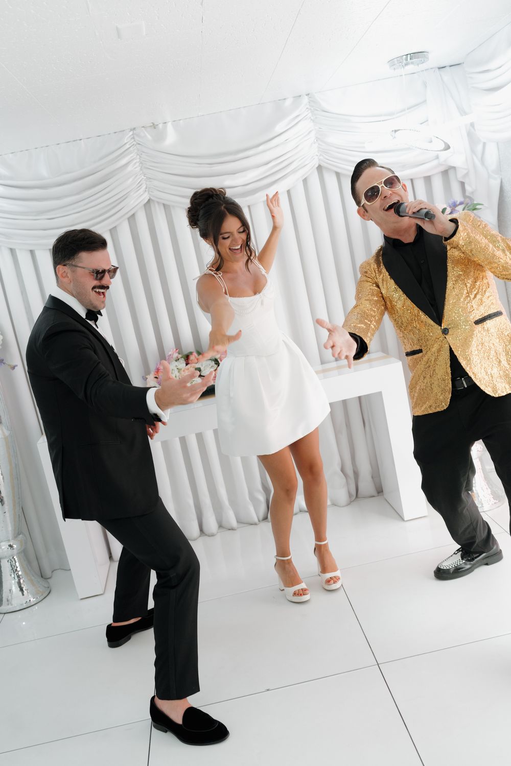 Bride in white mini dress dancing joyfully with groom in black tux and singer in gold jacket at wedding reception.