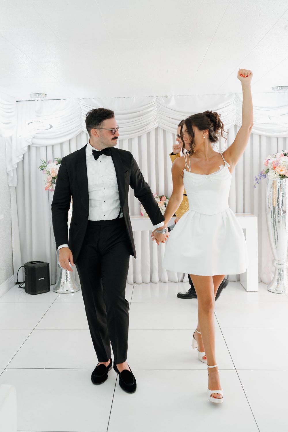 Joyful bride in short white dress and groom in tuxedo dancing together at elegant white wedding venue.