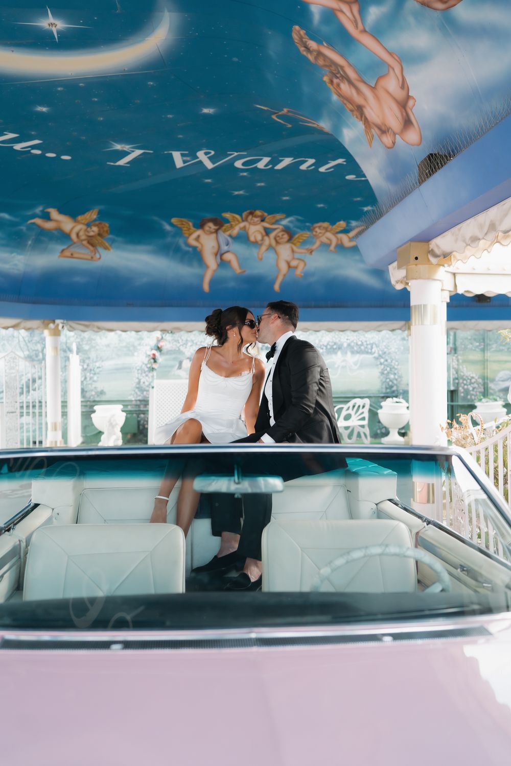 Couple in wedding attire kissing in a vintage pink convertible under a painted ceiling with cherubs.