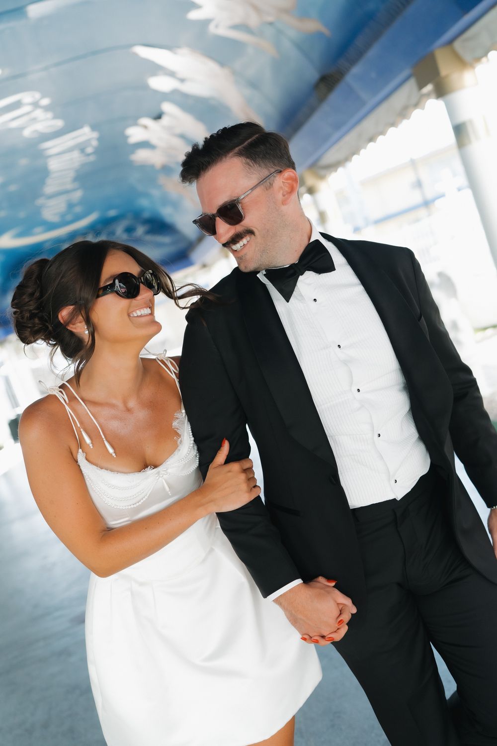 Smiling couple in sunglasses, woman in white dress and man in black tuxedo with bow tie, walking together outdoors.