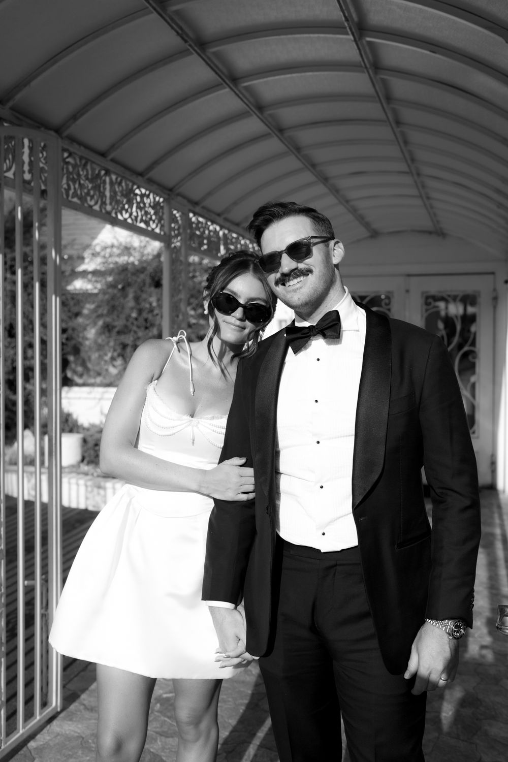 Black and white photo of an elegant couple in wedding attire, man in tuxedo and woman in white mini dress, both wearing sunglasses.