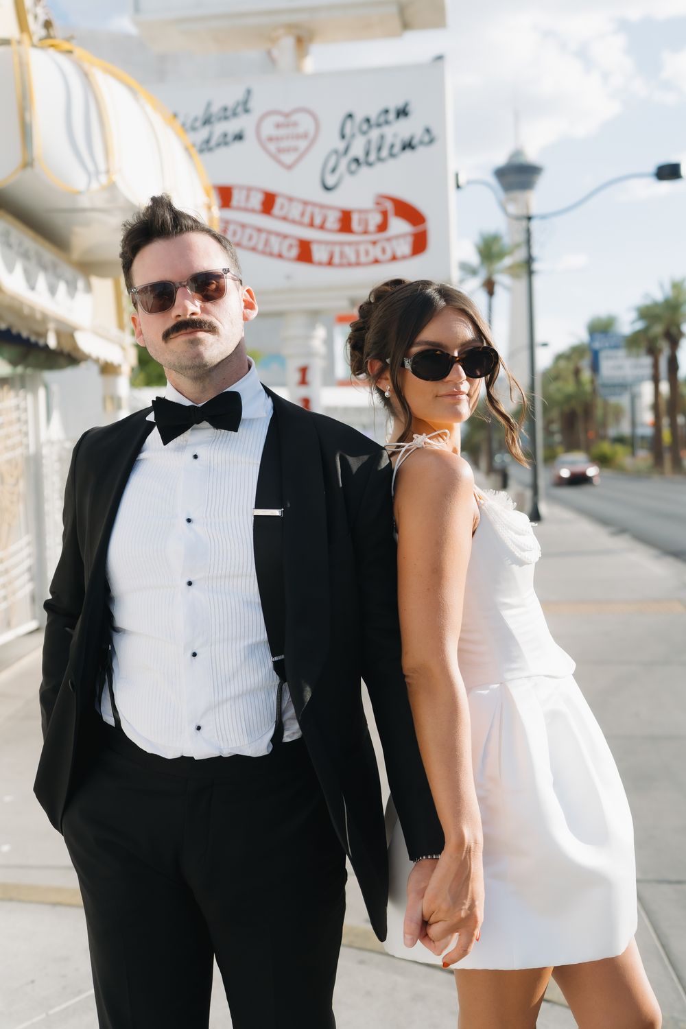Stylish newlyweds in tuxedo and white mini dress posing on Las Vegas Strip sidewalk.