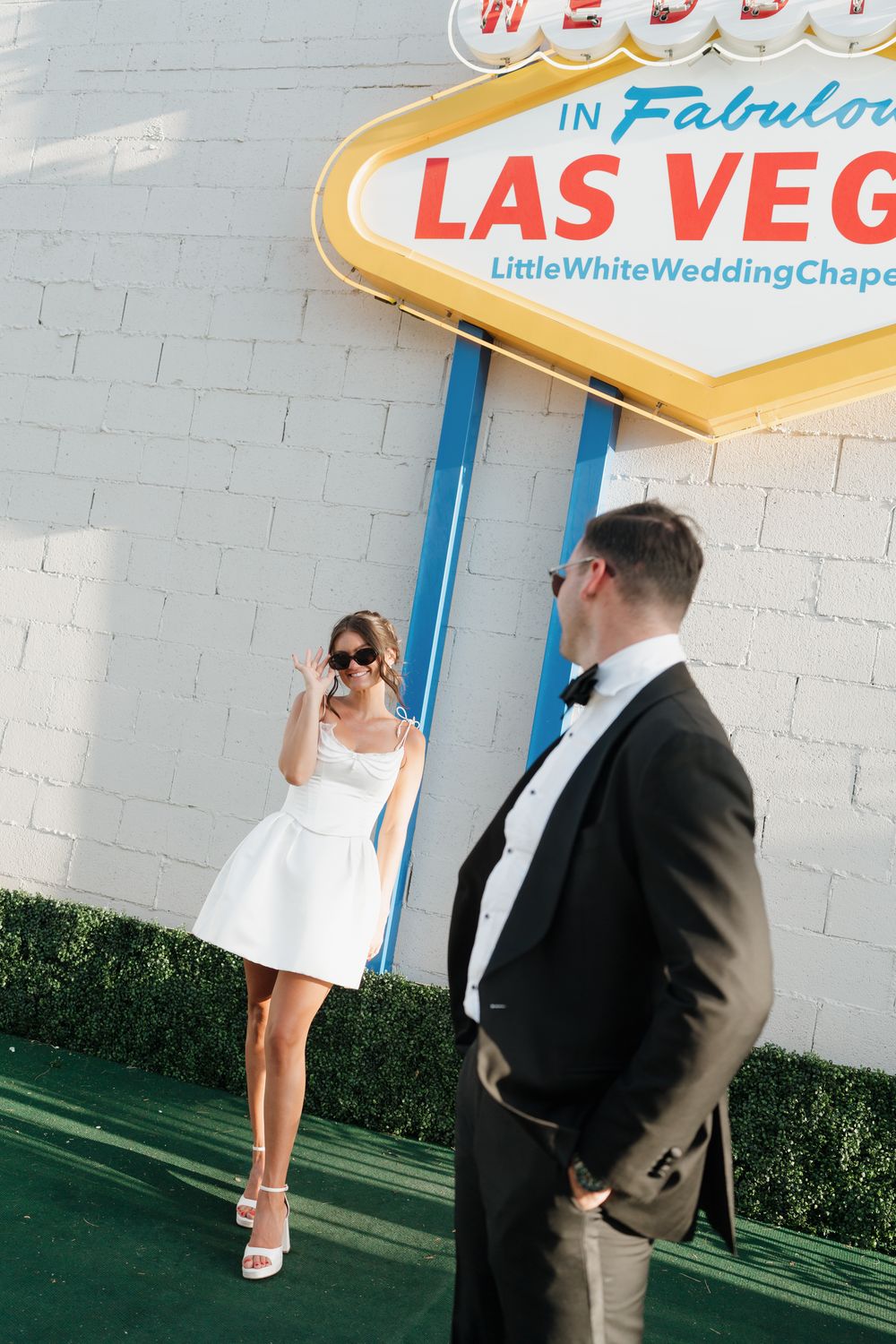 Bride in white mini dress poses with sunglasses in front of Las Vegas Little White Wedding Chapel sign.