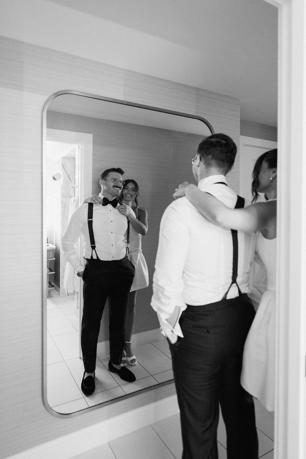 Black and white photo of a groom in suspenders and bow tie admiring his reflection in a mirror with bride nearby.