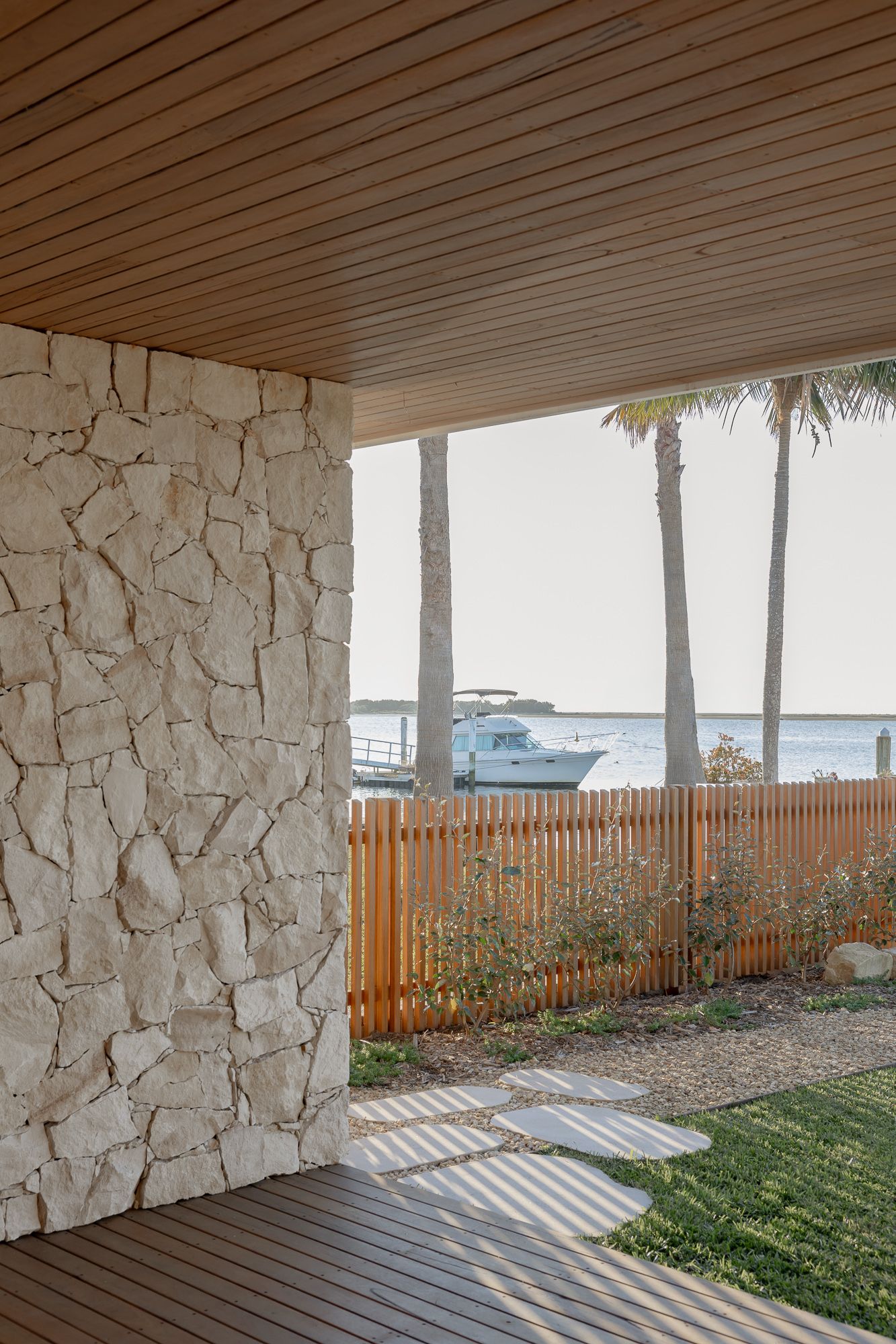 Natural stone wall beneath a timber-lined ceiling framing palm trees and the river, architectural photography on the NSW South Coast.