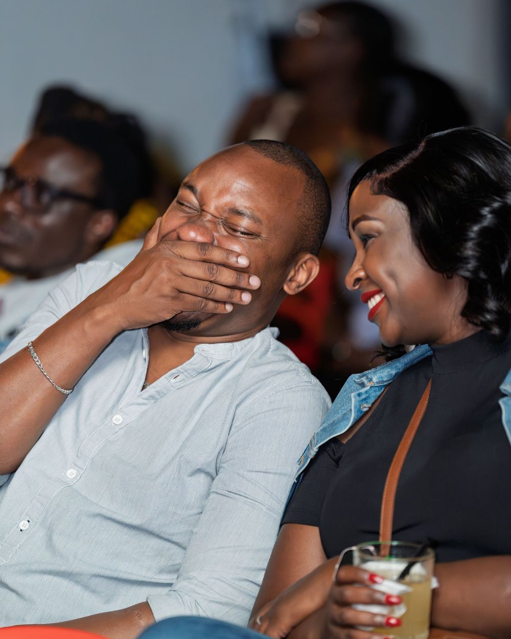 Couple laughing together at a comedy show, man covering his mouth while woman holds a drink.
