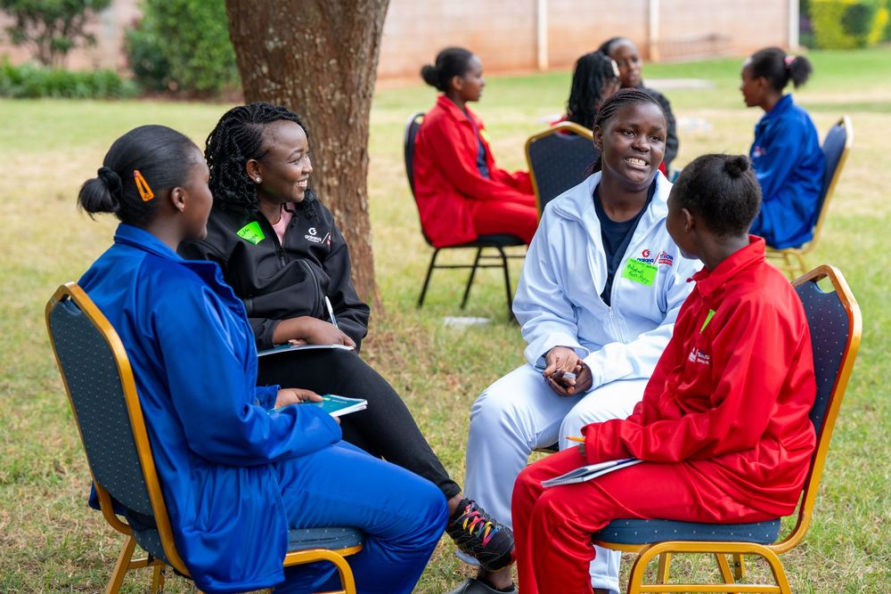 Young women in colorful tracksuits sit in a circle outdoors having a group discussion at a Kenyan foundation event.