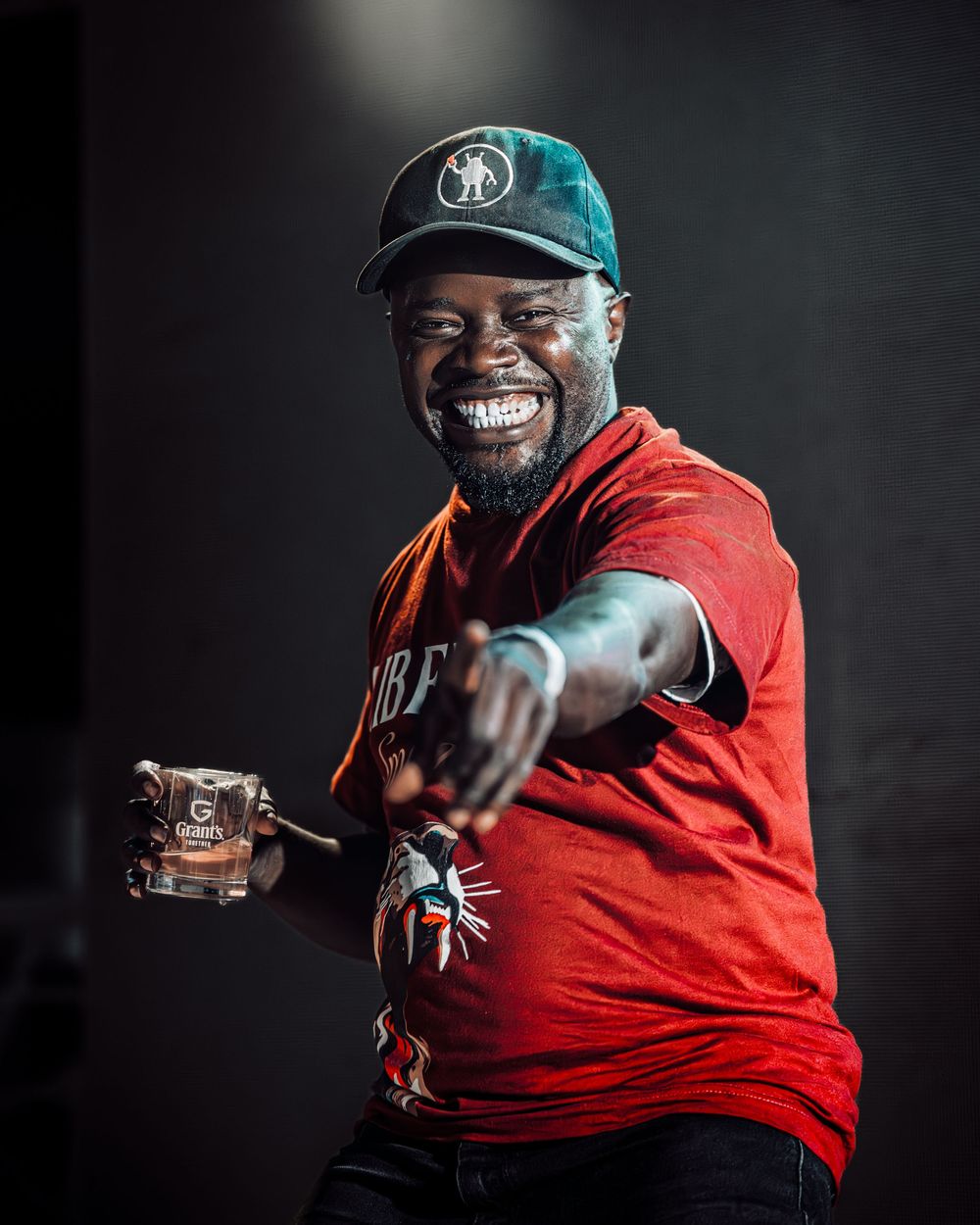 Smiling man in red shirt and cap pointing at camera holding a drink at an event.