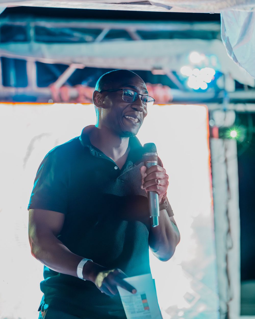 A smiling man in a dark polo shirt speaks into a microphone at a brightly lit outdoor event.