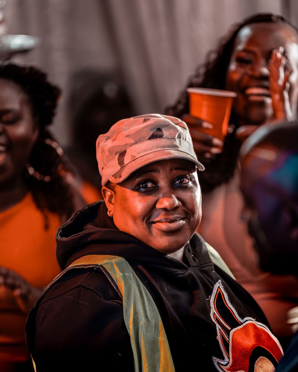 Smiling woman in camo cap and black hoodie at a lively outdoor evening social gathering.