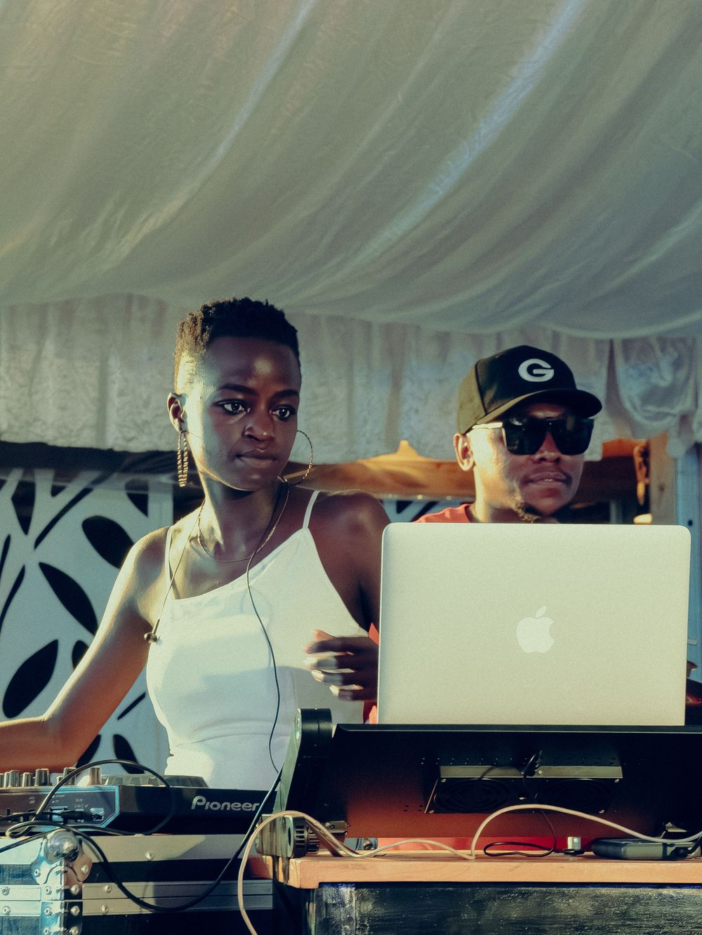 Female DJ performing at a Pioneer DJ setup with a man using a MacBook laptop at an outdoor music event.