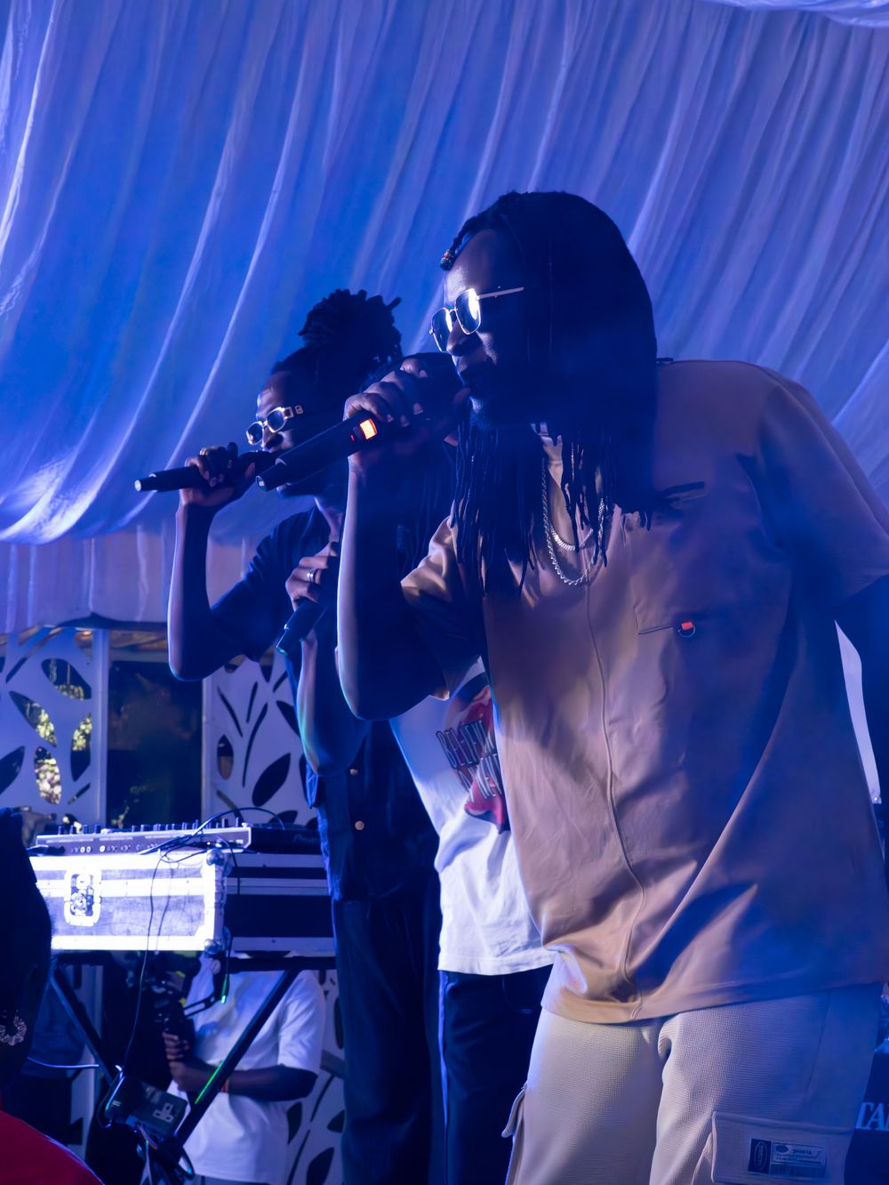 Two male performers singing into microphones on stage under blue lighting at a live music concert.