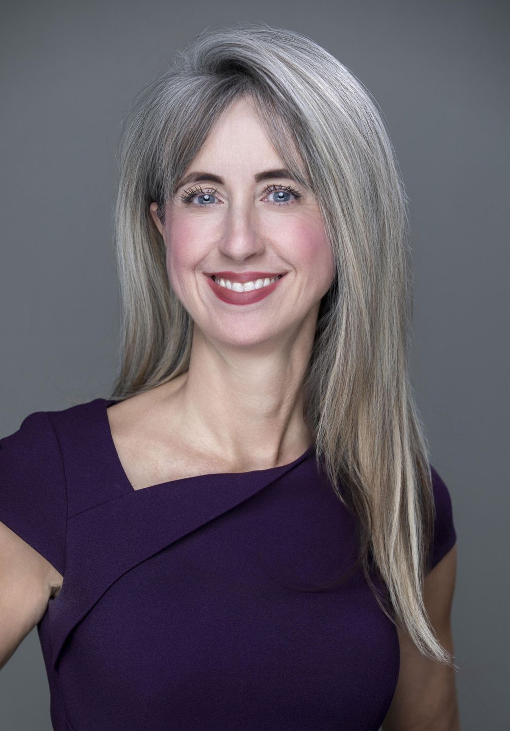 Professional headshot of a smiling woman with gray-streaked hair and blue eyes wearing a purple dress.