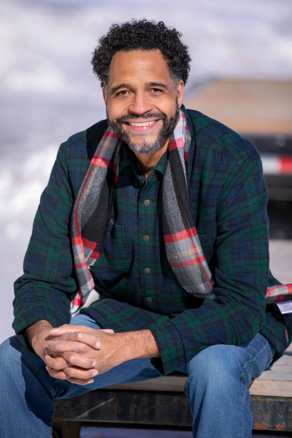 Smiling man with beard wearing plaid shirt and scarf sits outdoors in snowy setting.